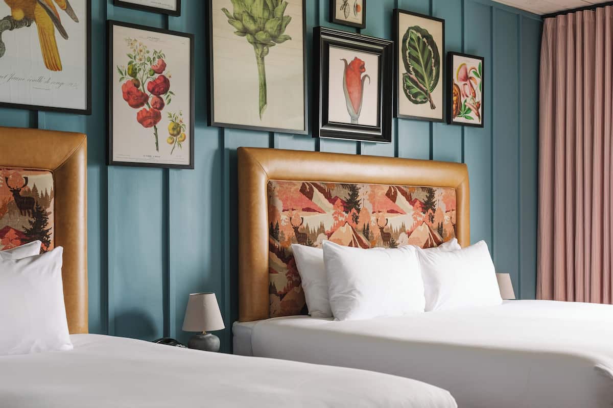 A hotel room with two beds, a headboard with a floral pattern, and framed botanical prints on the wall.
