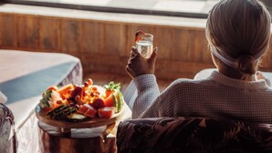 Una persona sentada junto a una ventana con vista a un patio, sosteniendo un vaso de bebida, y una mesa de madera con una bandeja de frutas al frente.