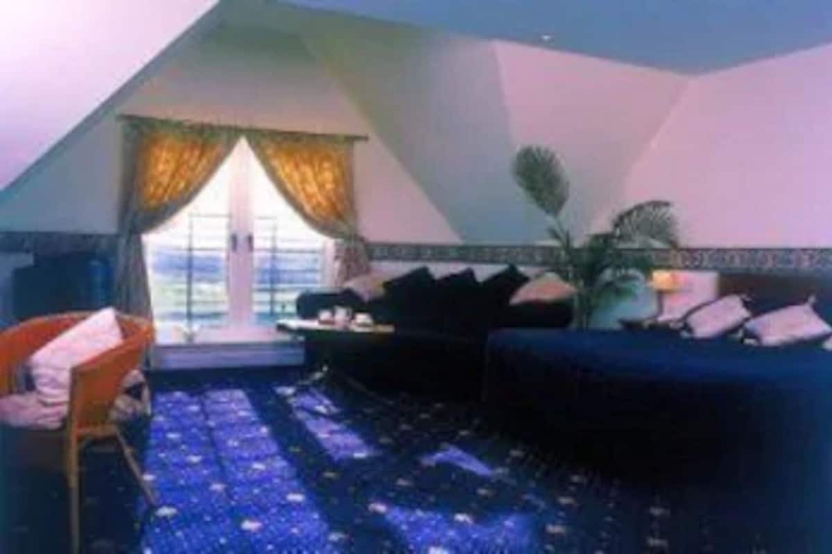 A room with a sloped ceiling, a large window with curtains, a dark blue sofa, a small table, and a chair.
