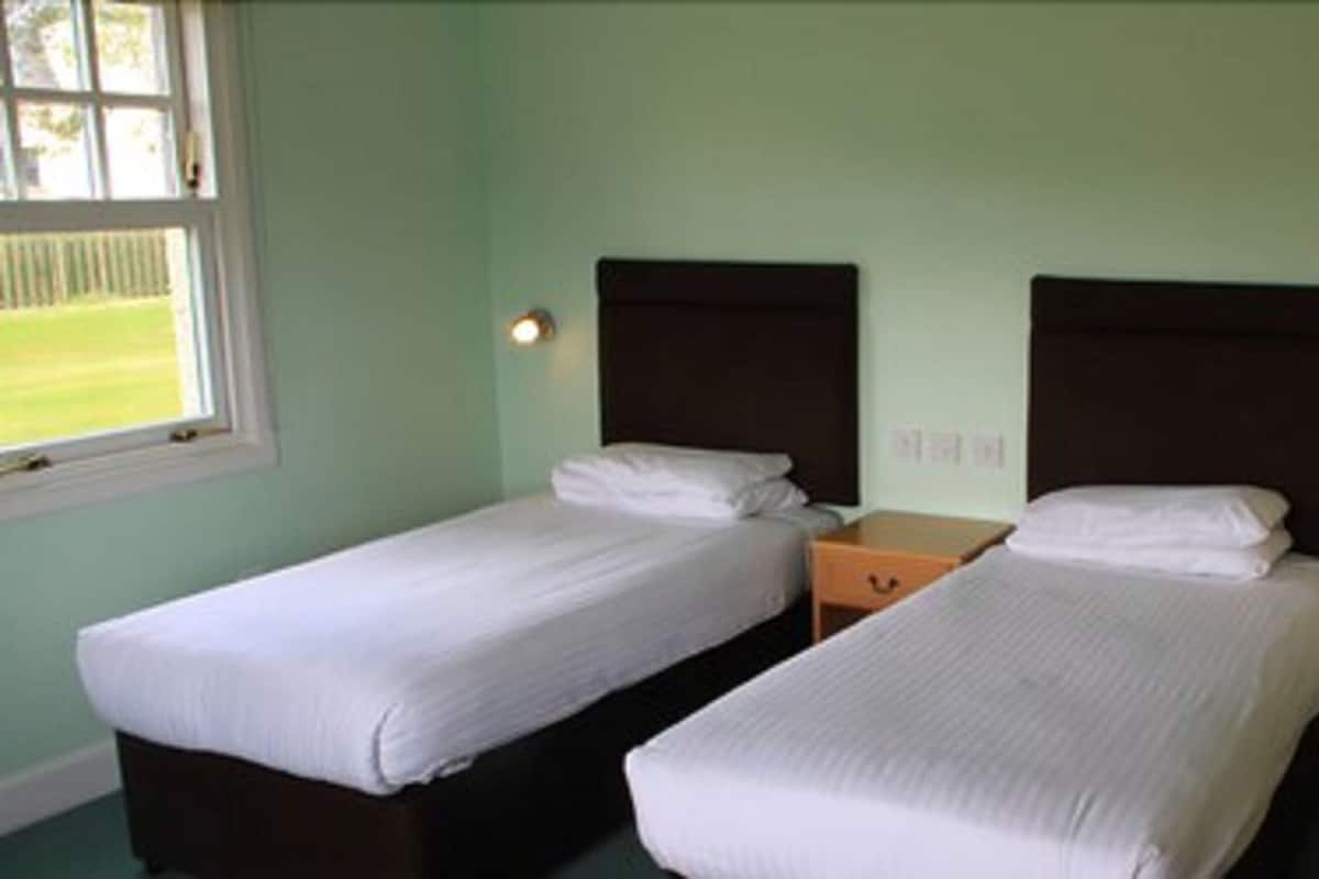 A room with two single beds, a small wooden nightstand, and a window with a view of a green lawn.