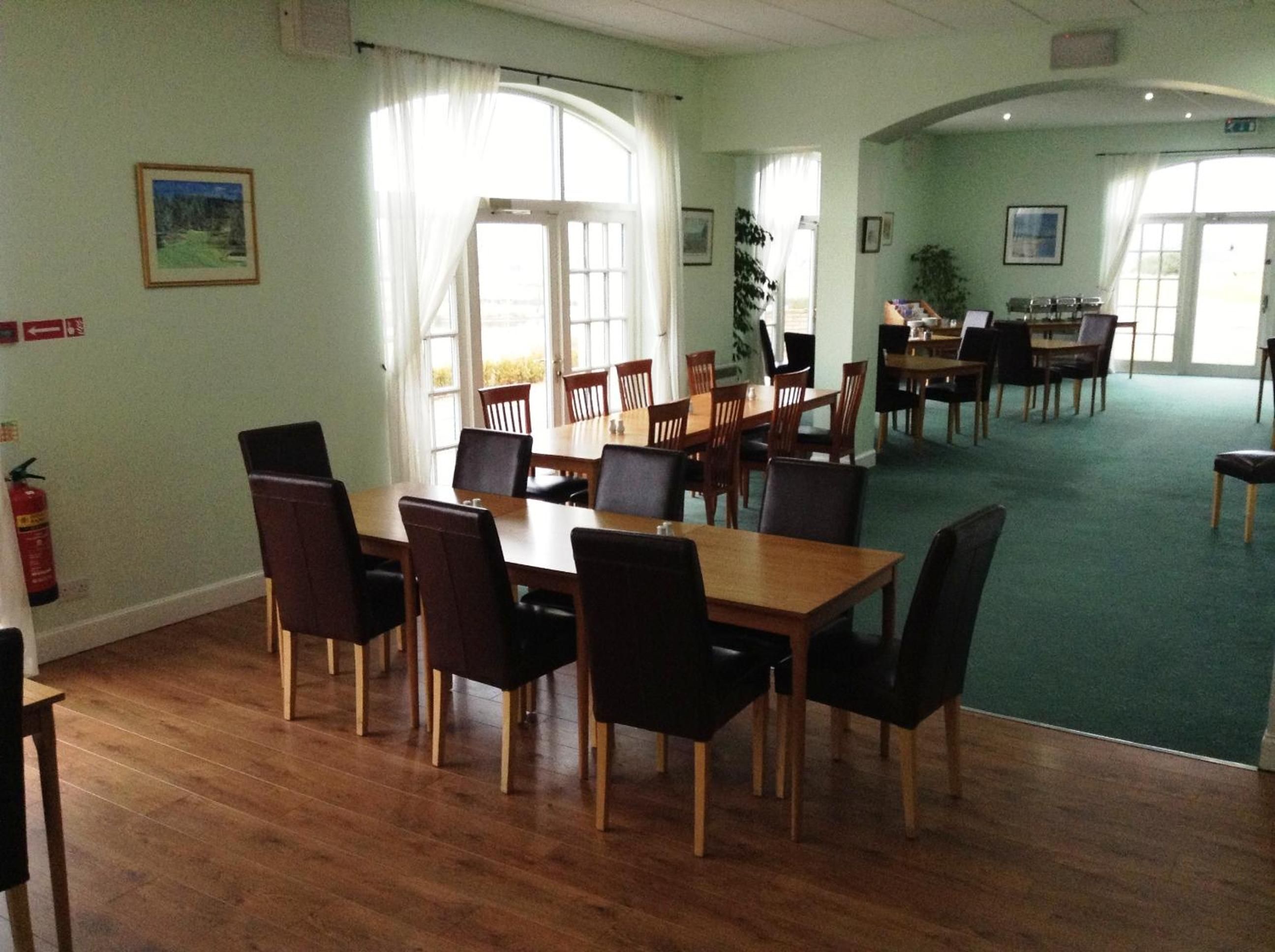 A dining area with wooden tables and chairs, large windows with curtains, and a green carpeted floor.