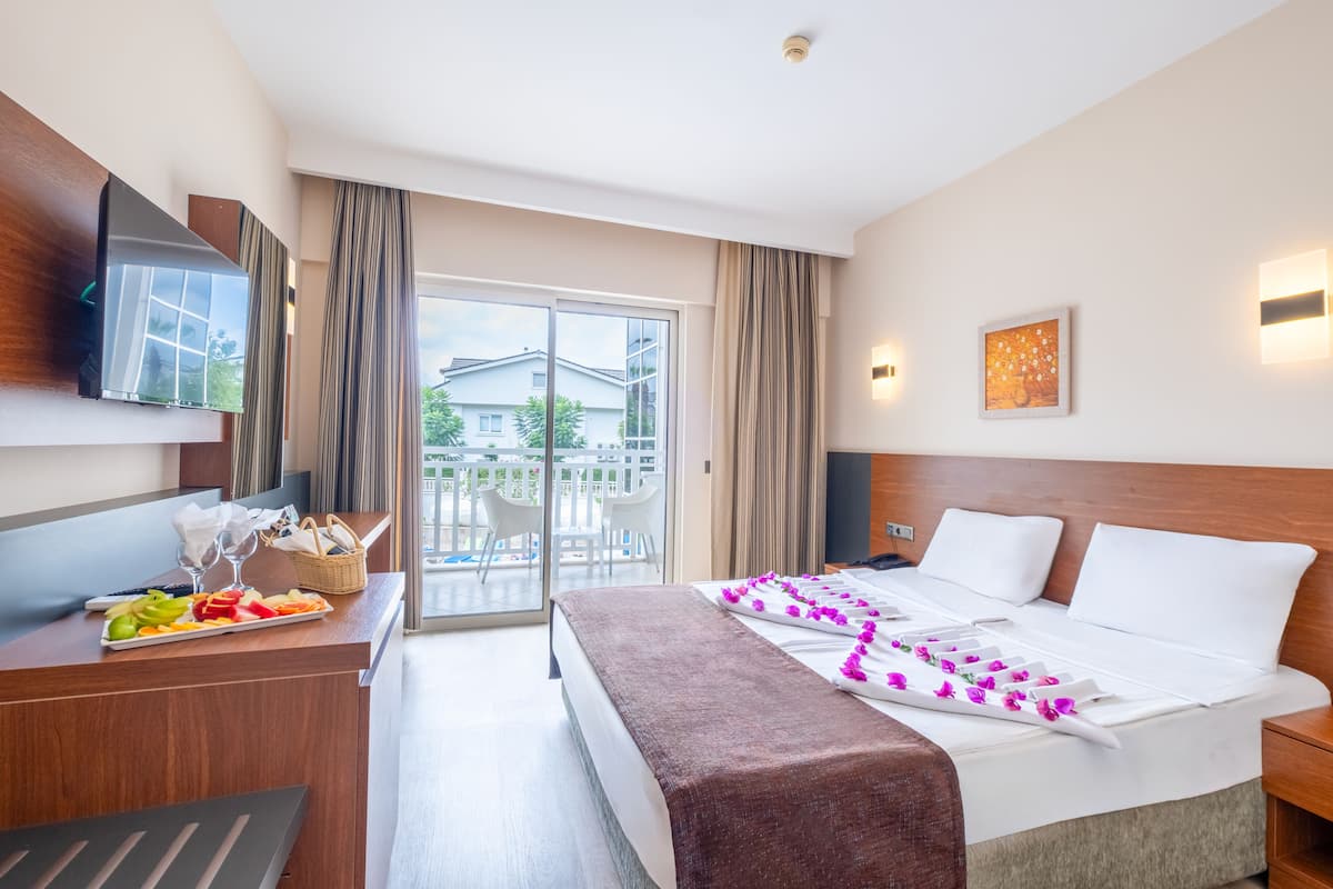 Ein Hotelzimmer mit Bett, Schreibtisch mit Obstteller, Fernseher und Balkon mit Blick auf Gebäude.