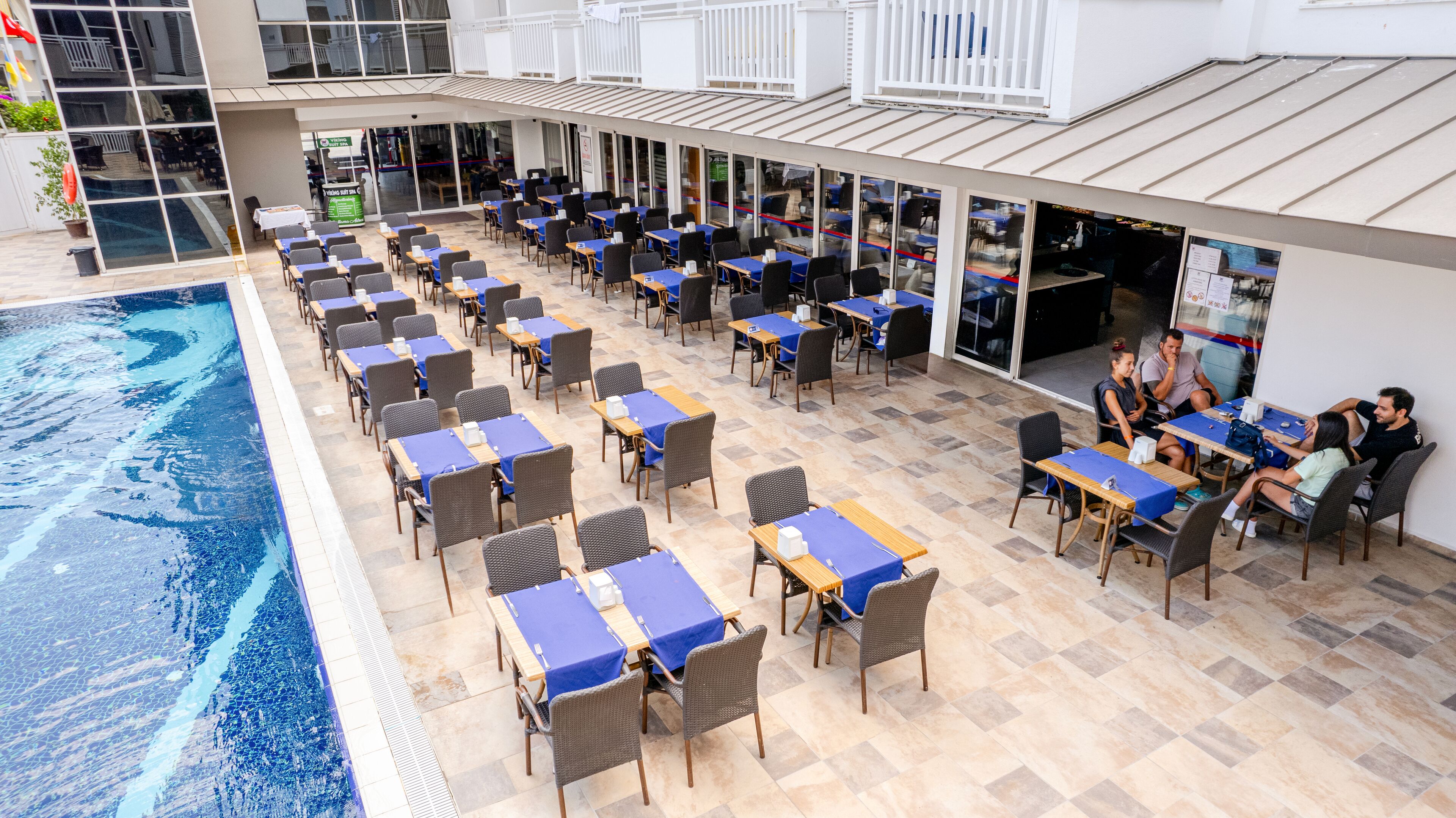 An outdoor dining area with tables and chairs, a pool, and a building with large windows and a balcony.