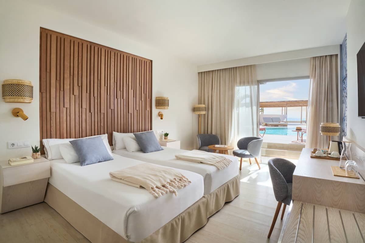 A hotel room with two beds, a desk, a chair, a small table, and a view of the pool.