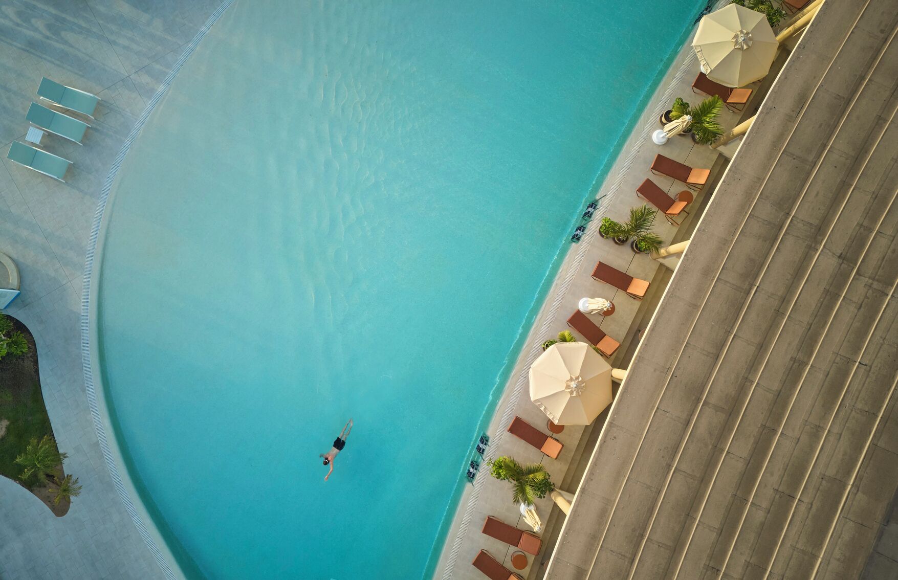 Vista aérea de una piscina con una persona nadando, sillas de descanso y sombrillas a su alrededor.