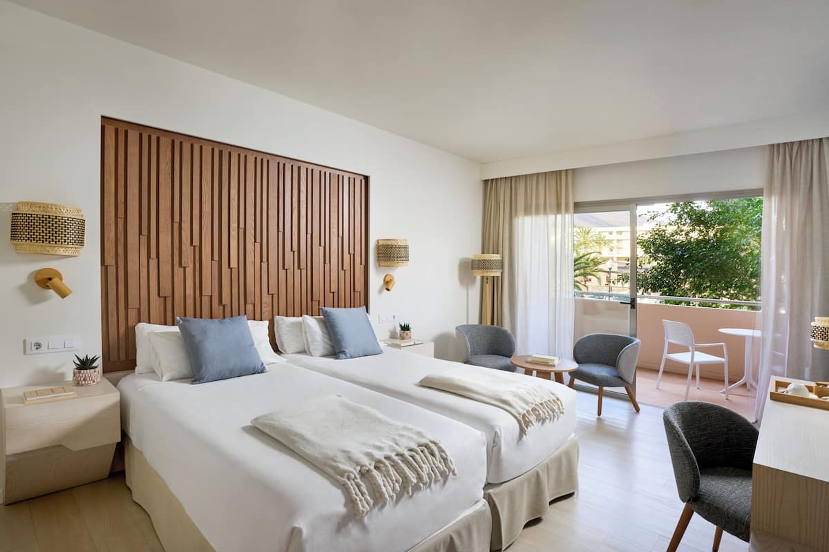 A hotel room with two beds, a wooden headboard, a balcony with a view, and a small seating area.
