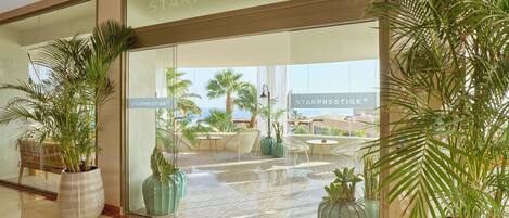 A hotel lobby with a large glass facade, potted plants, and a view of palm trees and a pool.