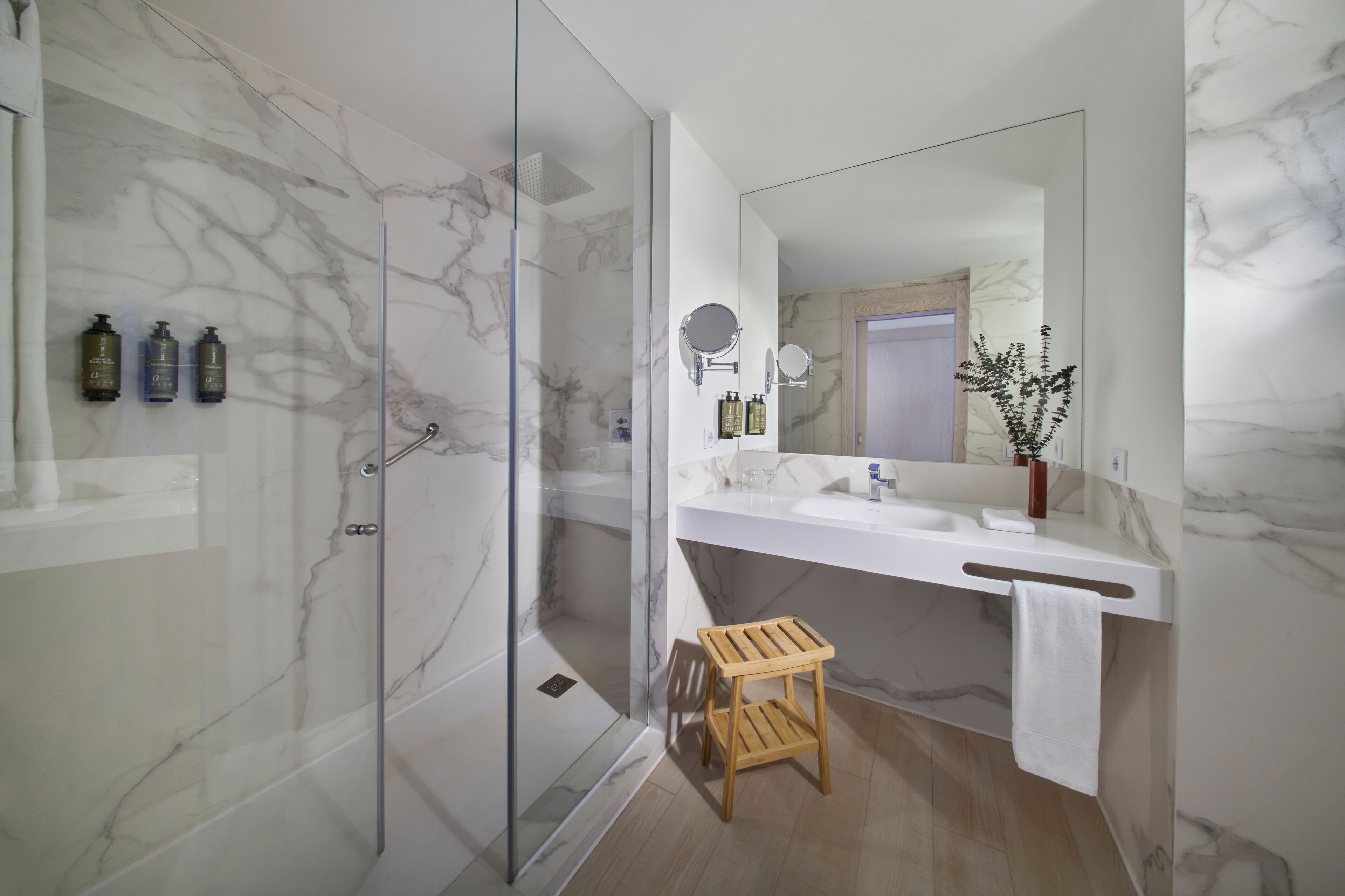 Un baño moderno con ducha de vidrio, un taburete de madera, un espejo grande y paredes de mármol.