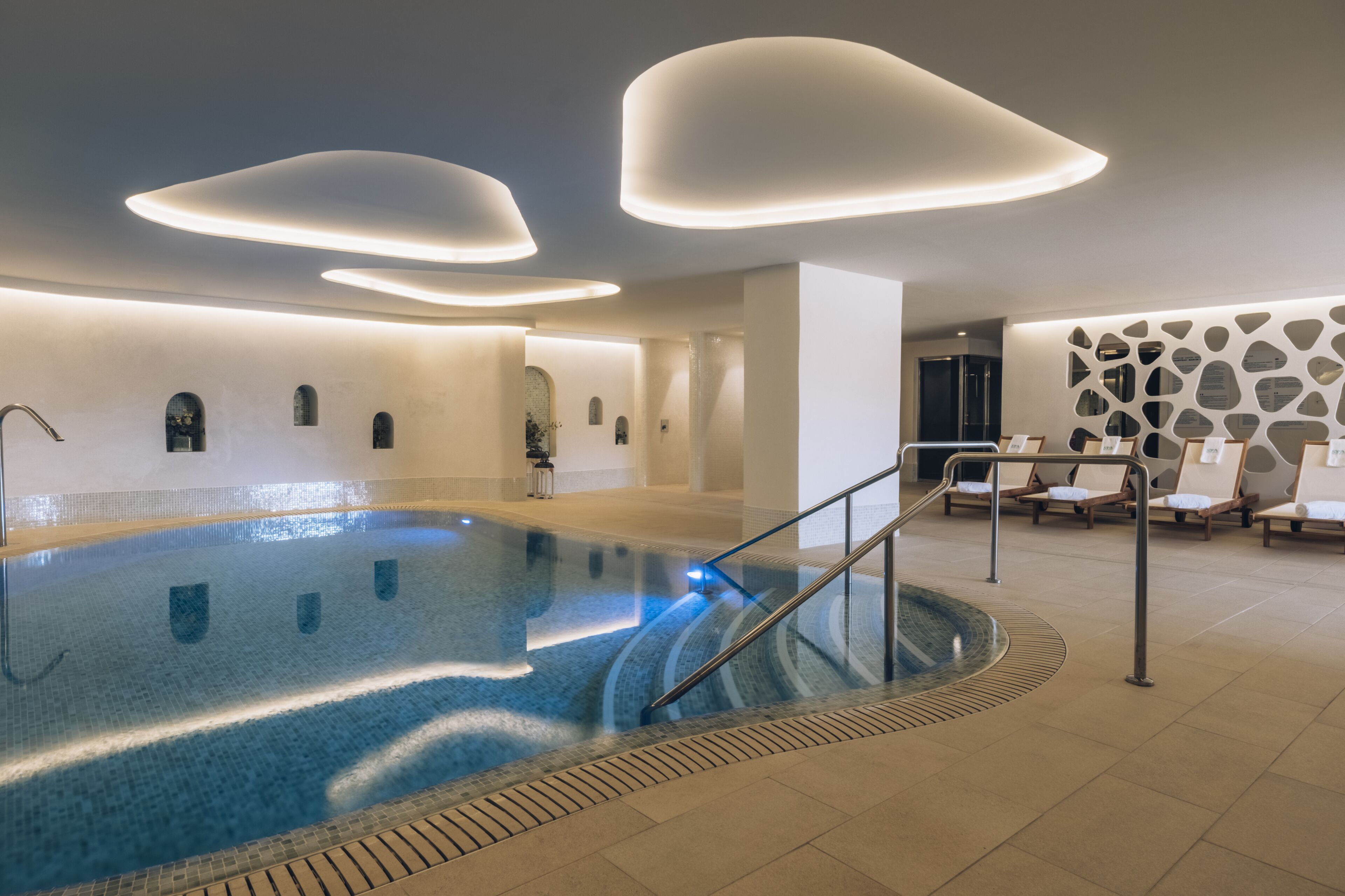 A modern indoor swimming pool with a tiled floor, a central water feature, and a seating area with lounge chairs.