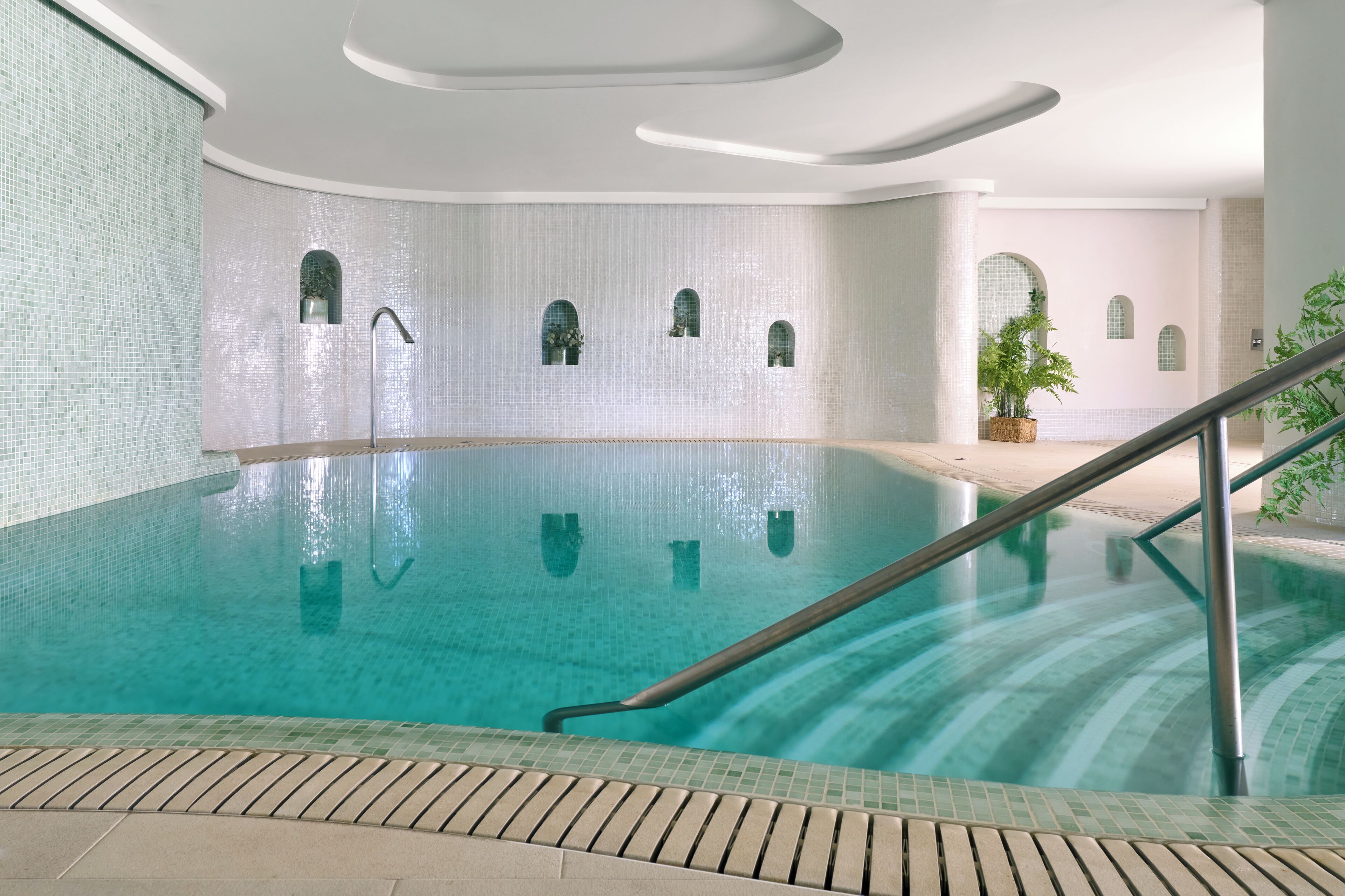 Piscina interna com água cristalina, corrimão de metal e paredes revestidas com janelas em arco.