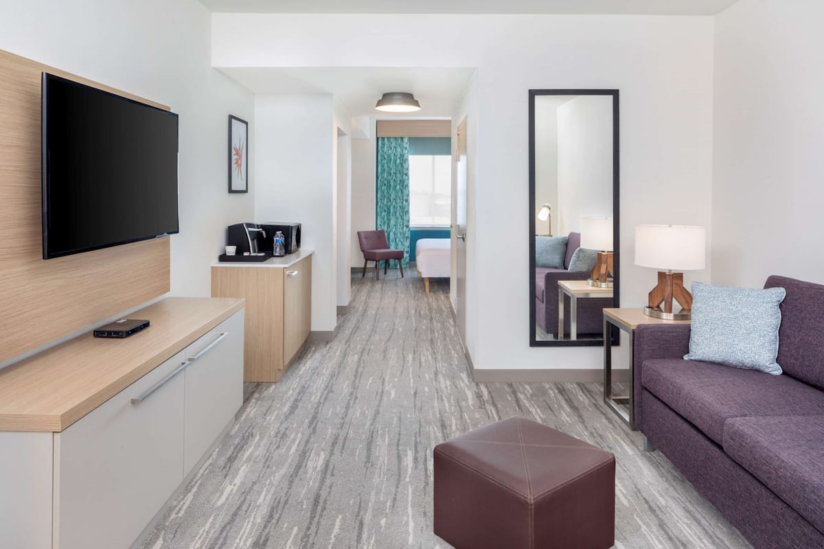 A modern hotel room with a flat-screen TV, a sofa, a coffee table, a desk with a computer, and a mirror.