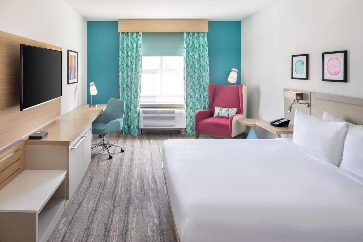 A hotel room with a bed, desk, chair, TV, air conditioning unit, and curtains.