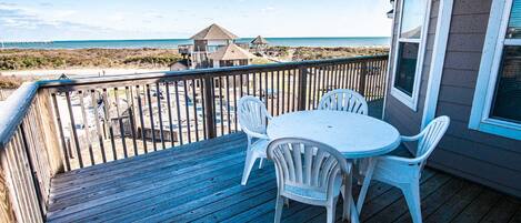 A wooden deck with a white table and chairs, overlooking a beach and ocean.