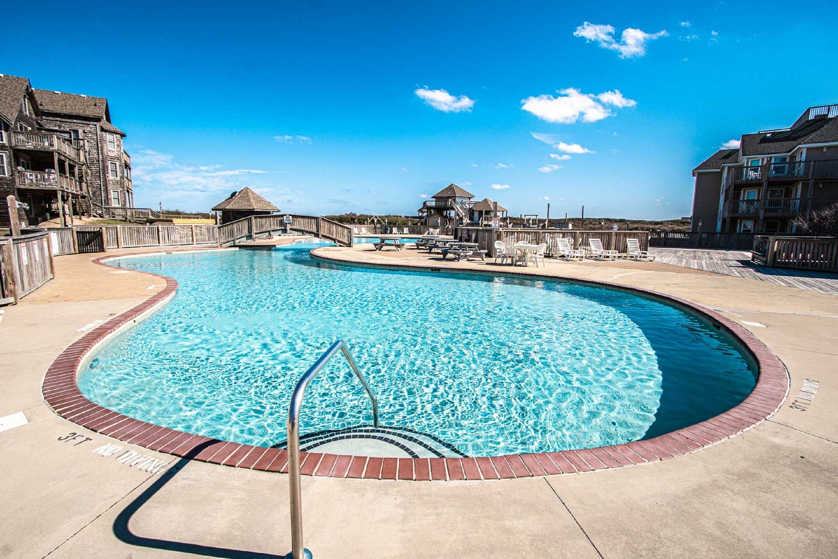 Ein großer, gebogener Pool mit einer Holzterrasse und Liegestühlen, umgeben von Gebäuden mit Balkonen und blauem Himmel.