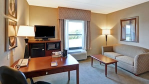 A hotel room with a desk, a television, a sofa, a lamp, and a window with curtains.