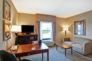 A hotel room with a desk, a television, a sofa, a lamp, and a window with curtains.