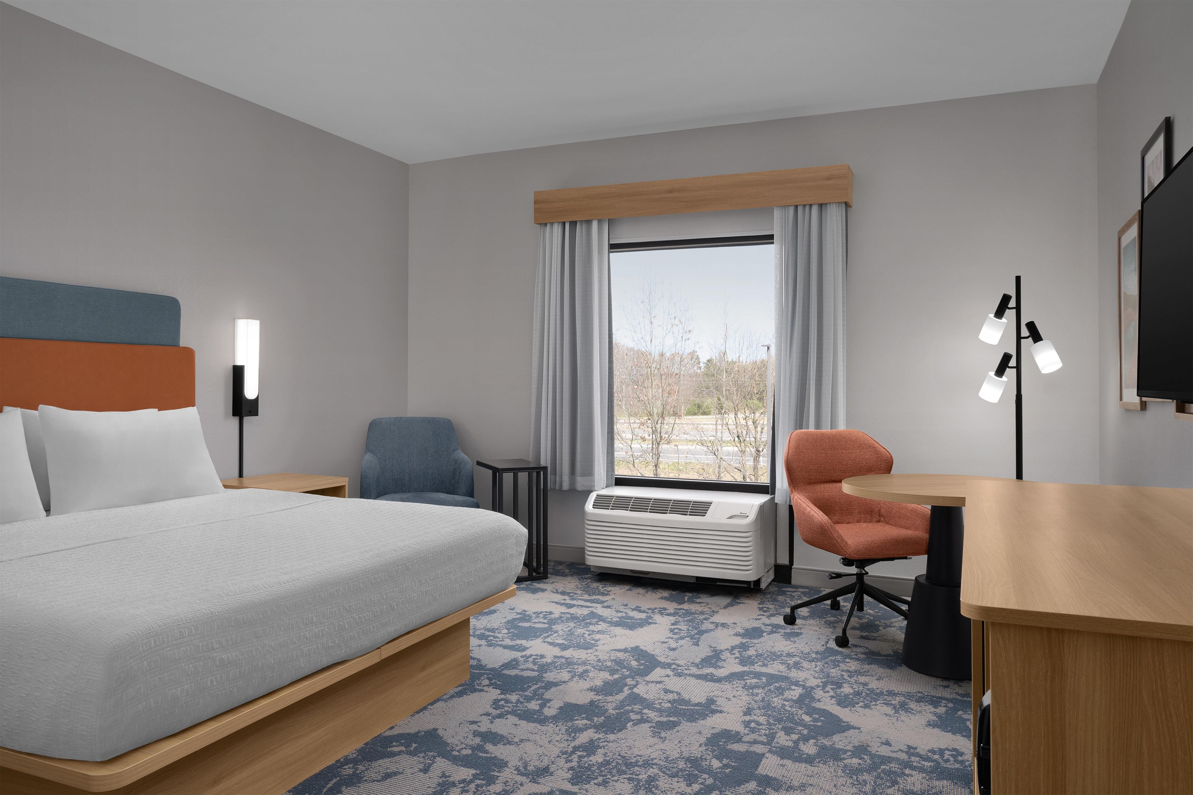 A hotel room with a bed, a desk, a chair, a window with curtains, and a wall-mounted TV.
