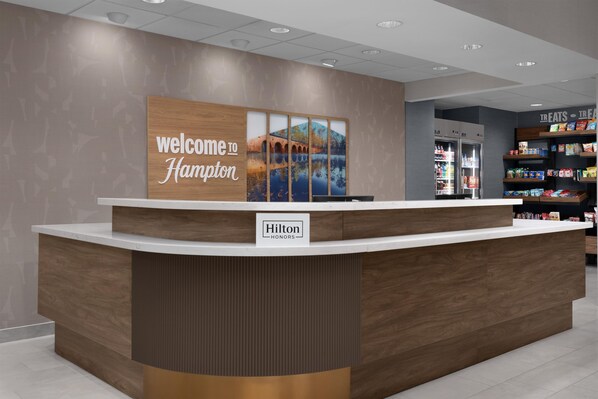 Una recepción de hotel con un letrero que dice 'Welcome to Hampton', una insignia de 'Hilton Honors' y un área de exhibición de alimentos al fondo.