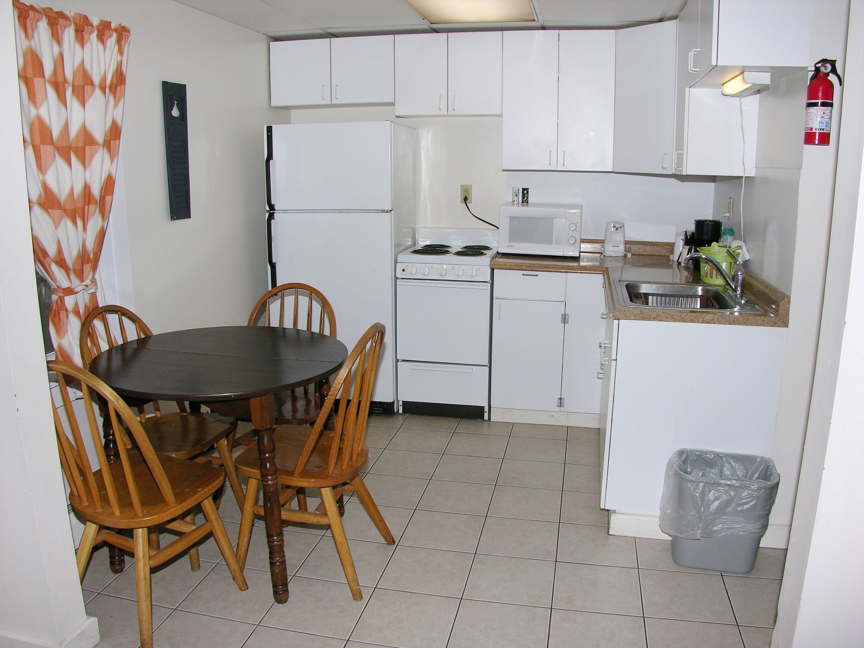 Une cuisine compacte avec des meubles blancs, une cuisinière, un four à micro-ondes et un évier. On y trouve une table à manger avec des chaises et une poubelle.