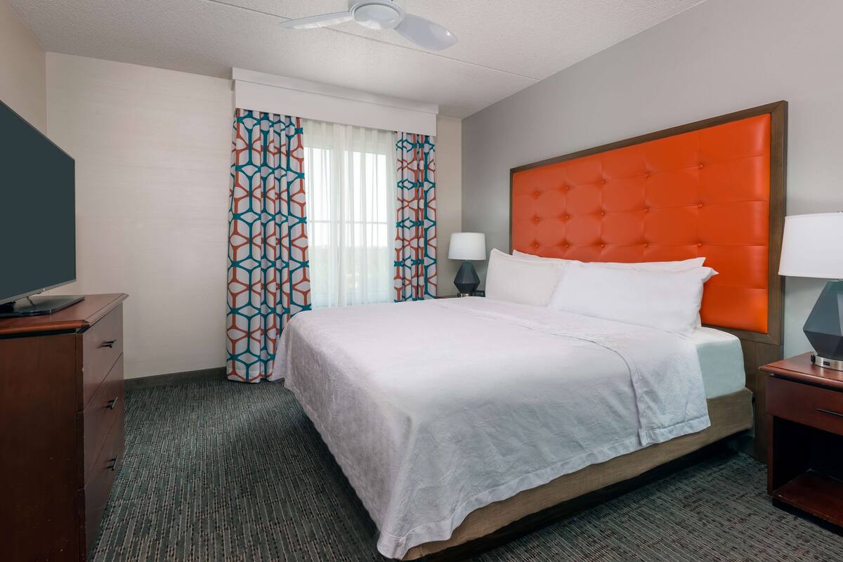 Una habitación de hotel con cama, mesita de noche, escritorio y ventana con cortinas estampadas.