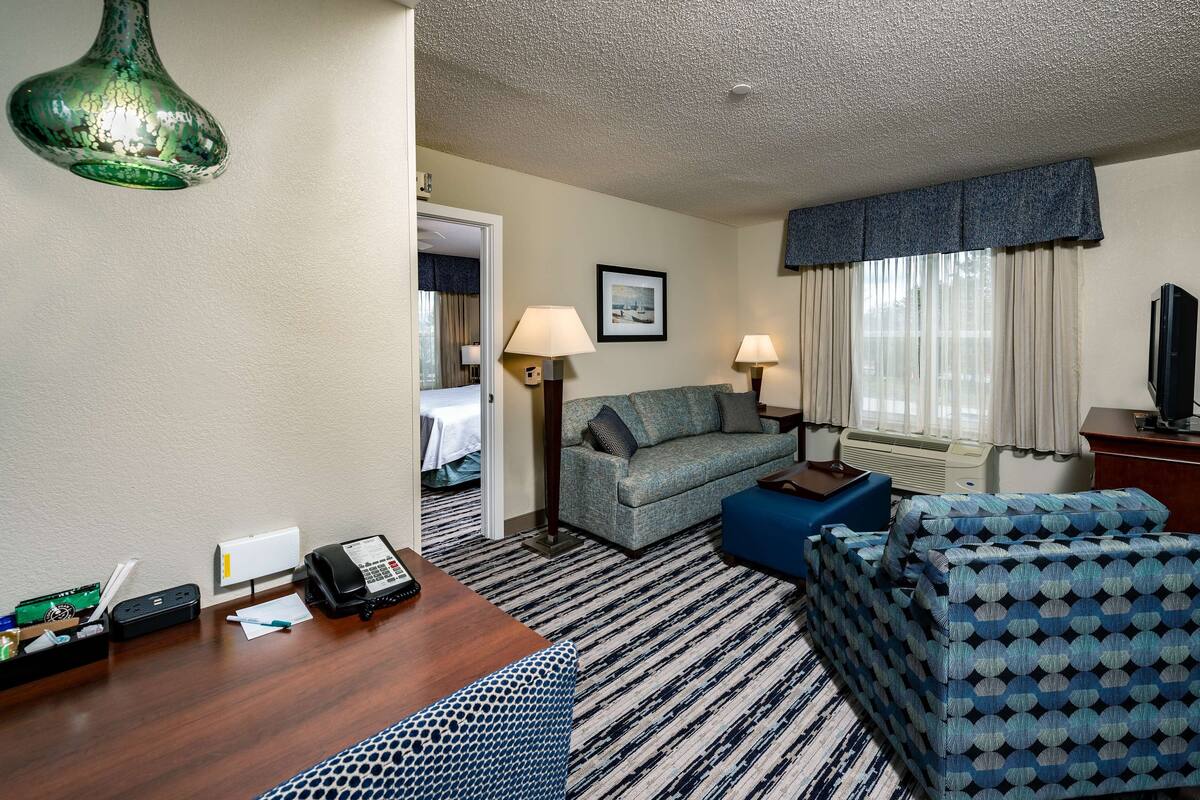 A hotel room with a sofa, a blue patterned sofa, a wooden desk with a phone and a lamp, a wall-mounted green light fixture, a framed picture, and a window with curtains.