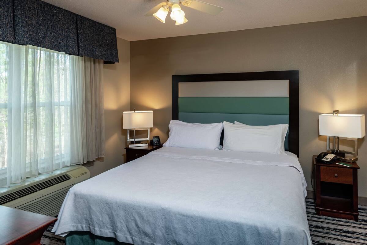 A hotel room with a large bed, two bedside tables with lamps, a ceiling fan, and a window with sheer curtains.