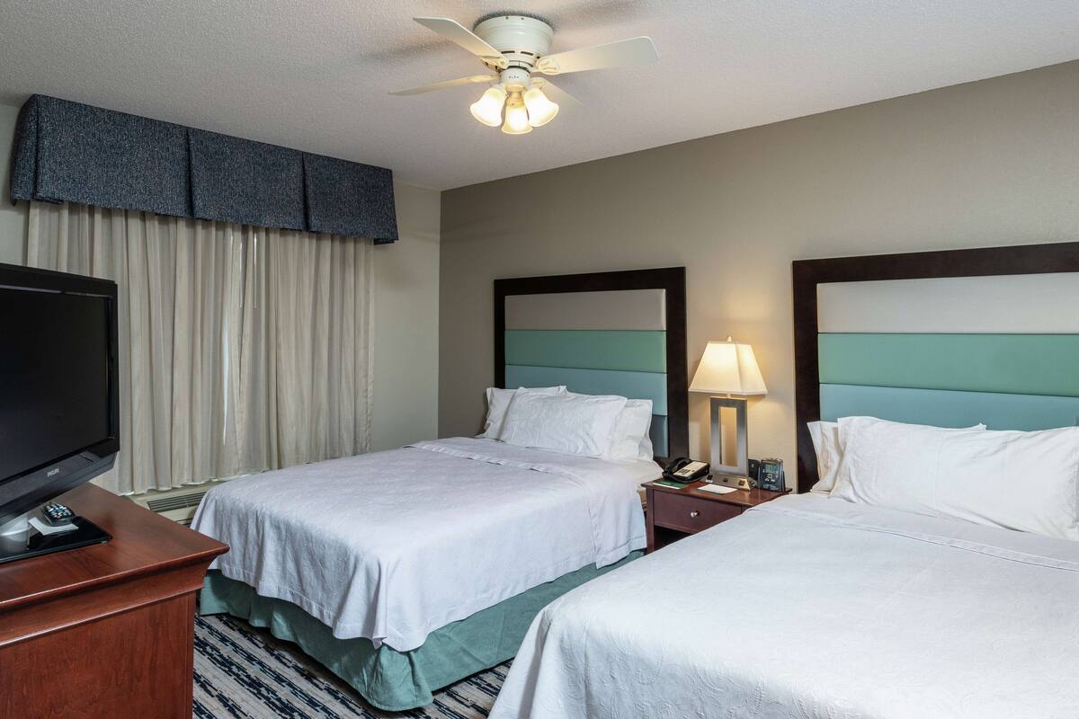 A hotel room with two beds, a television, a ceiling fan, and a nightstand with a lamp.