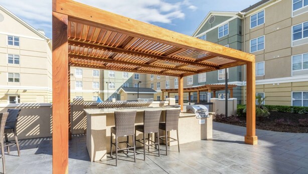 Un patio exterior con una pérgola de madera, una zona de bar con taburetes y una parrilla.