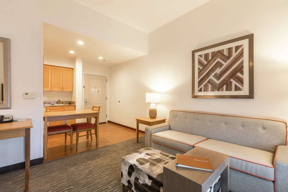 A modern hotel room with a sofa, a small table, a lamp, and a kitchen area in the background.