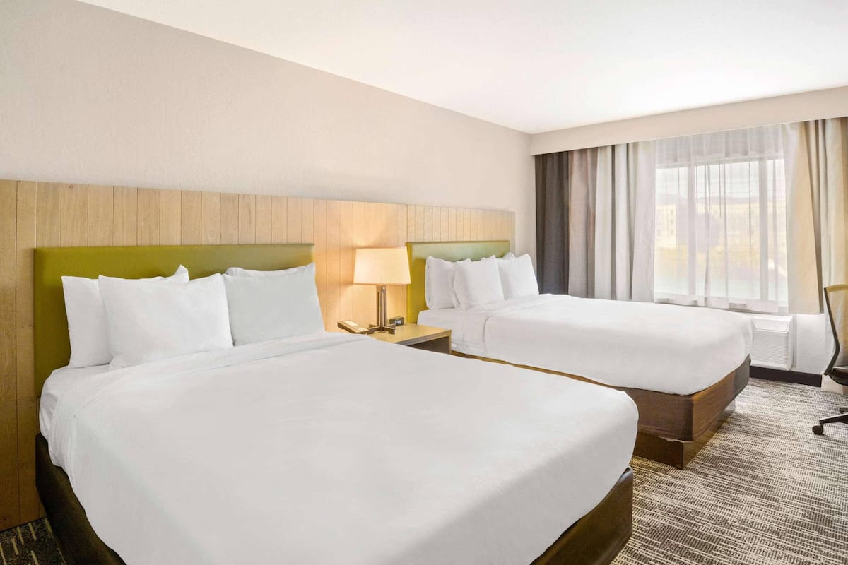 Habitación de hotel con dos camas, una mesita de noche con lámpara, una silla y un gran ventanal con cortinas.