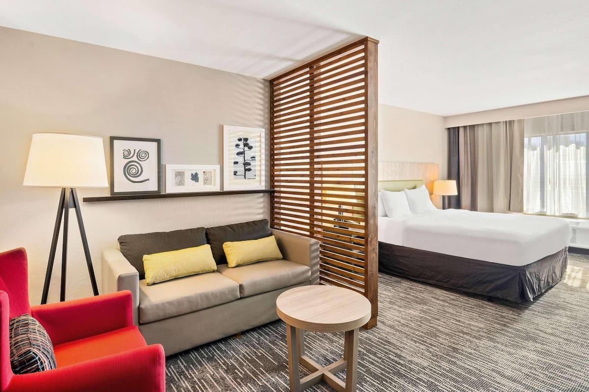 Una habitación de hotel con cama, sofá, sillón rojo, una pequeña mesa redonda y una lámpara de pie.