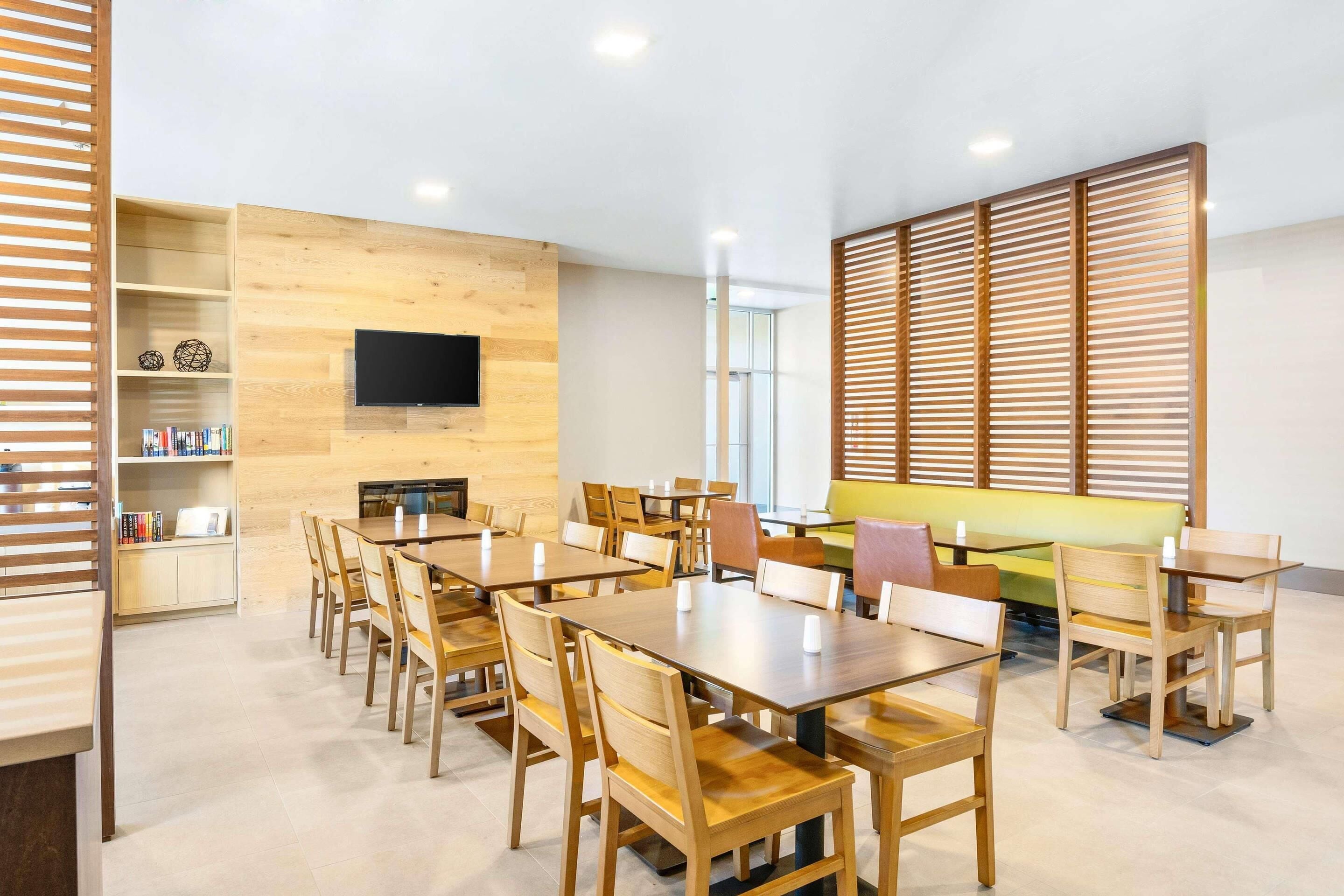 Un vaste espace de salle à manger avec des tables et des chaises en bois, un téléviseur à écran plat fixé au mur et une grande fenêtre laissant entrer la lumière naturelle.