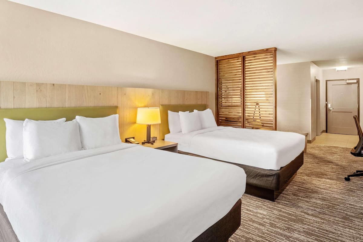 Habitación de hotel con dos camas, cabecero de madera, mesita de noche con lámpara y una puerta.