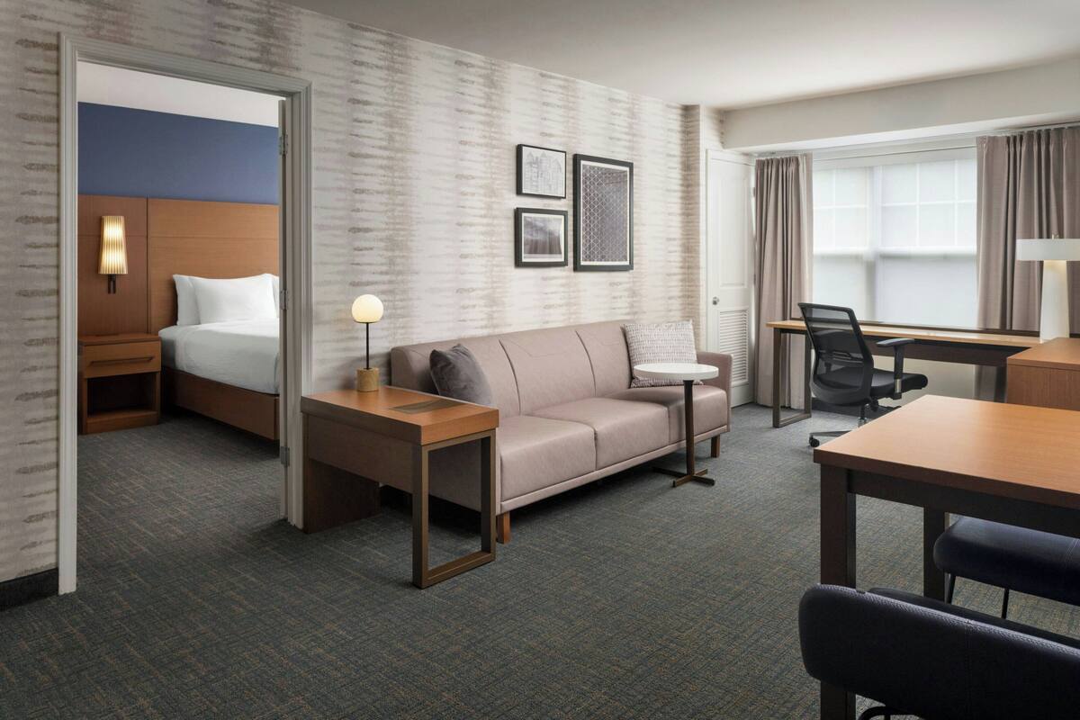 A hotel room with a bed, a sofa, a desk, and a chair. There are framed pictures on the wall and a lamp on the desk.