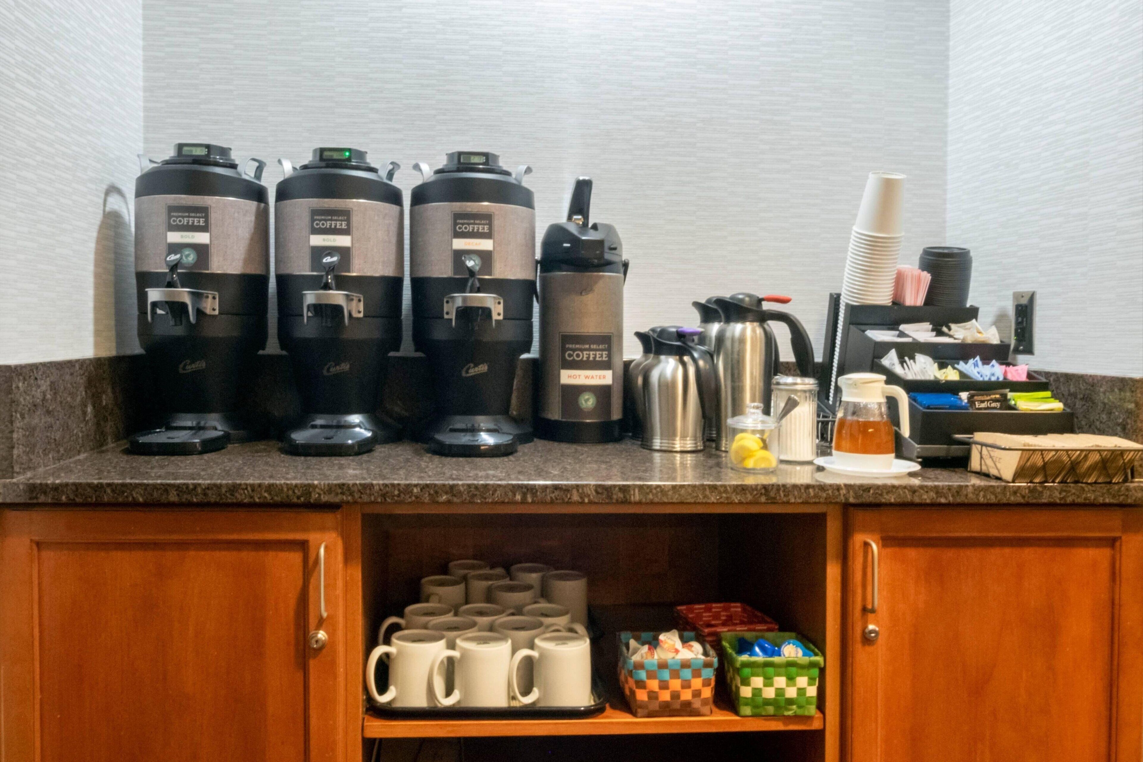 A coffee station with multiple coffee dispensers, a hot water dispenser, and a coffee pot. There are mugs and a pitcher on the counter.