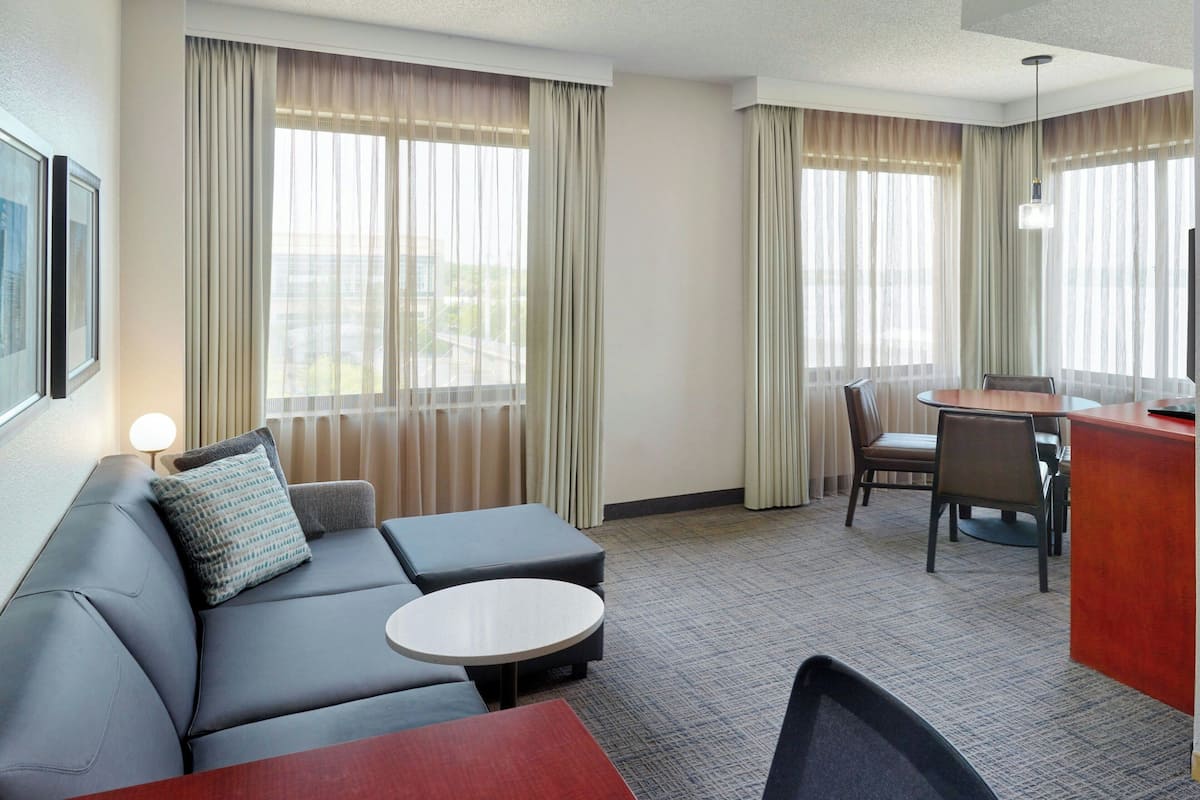 A hotel room with a sofa, a small round table, a dining table with chairs, and a view through large windows.
