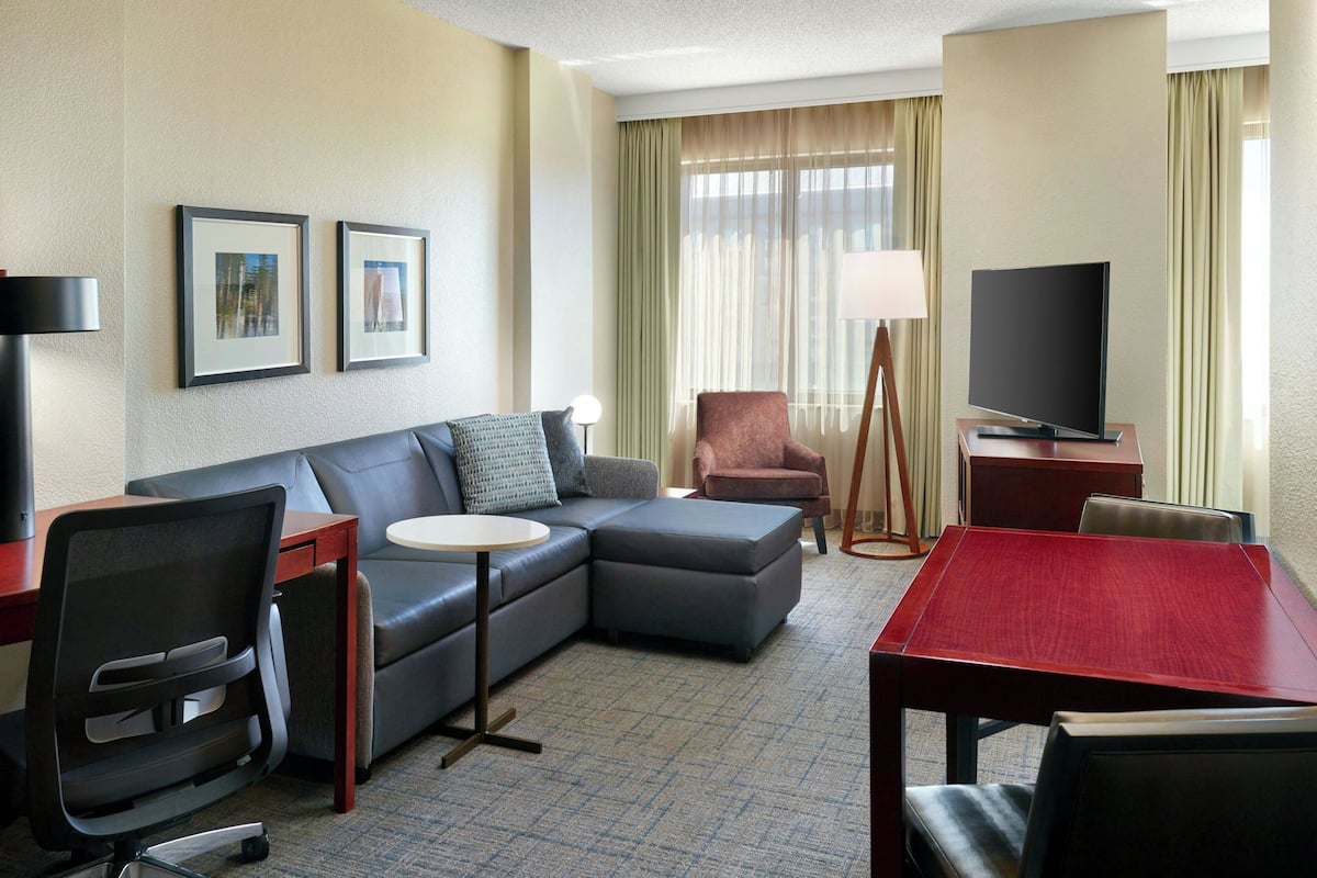 A hotel room with a sectional sofa, a small round table, a TV, a desk, and a chair.