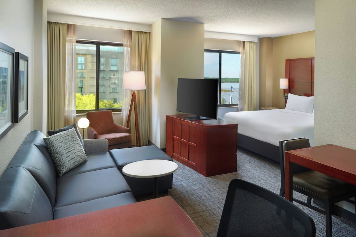 A hotel room with a bed, a sofa, a desk, a television, and a window with a view of buildings and greenery.