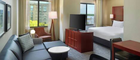 A hotel room with a bed, a sofa, a desk, a television, and a window with a view of buildings and greenery.
