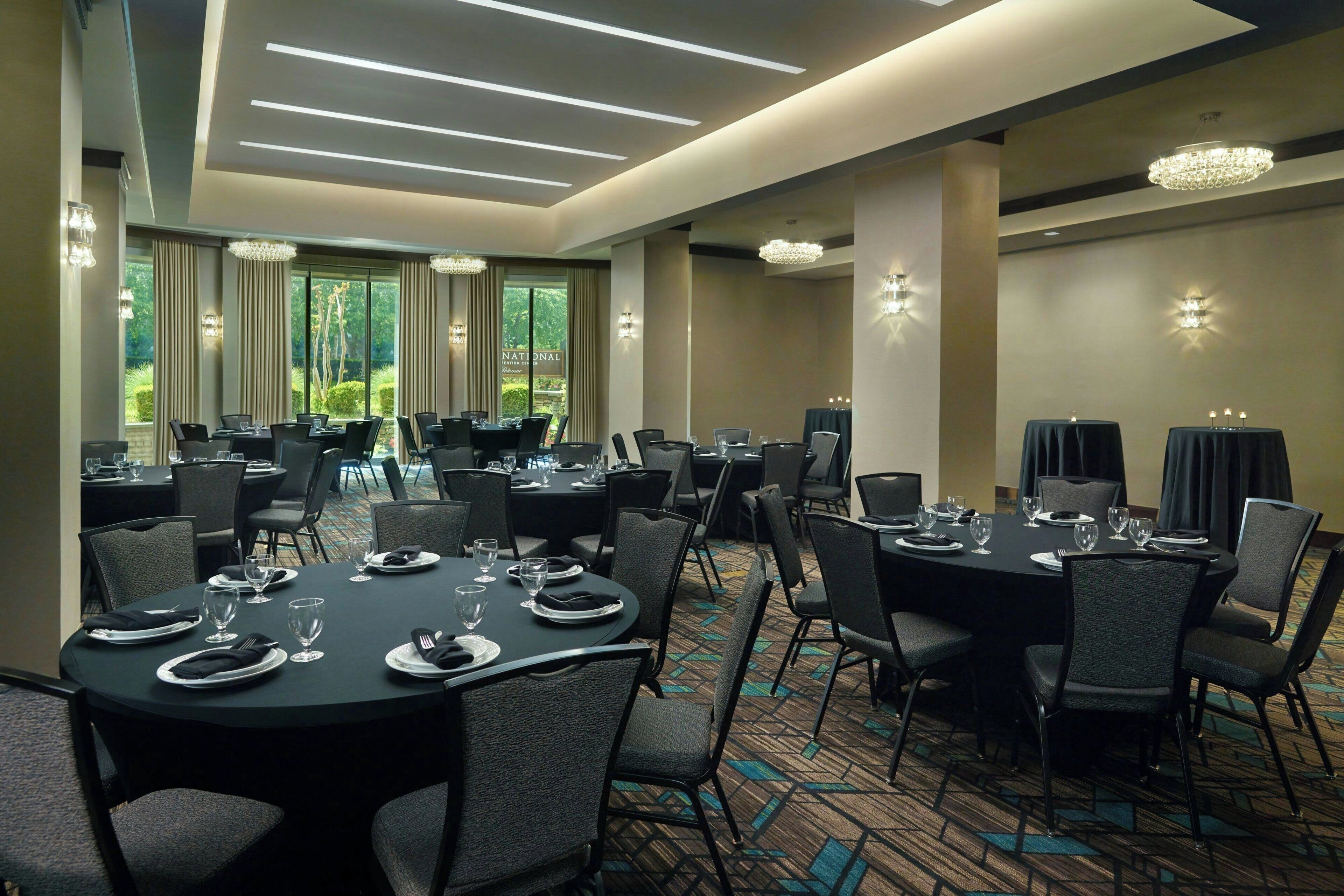 Un salón de conferencias con varias mesas dispuestas para comidas, ventanas amplias con cortinas y candelabros.