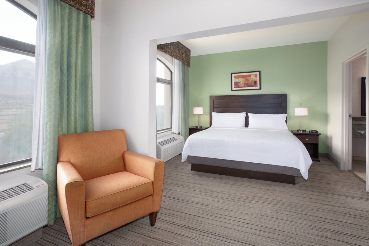 A hotel room with a bed, a chair, a window with curtains, a bedside table with a lamp, and a wall-mounted picture.