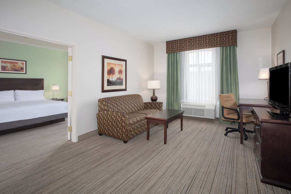 A hotel room with a bed, a sofa, a desk, a chair, a TV, and a window with curtains.