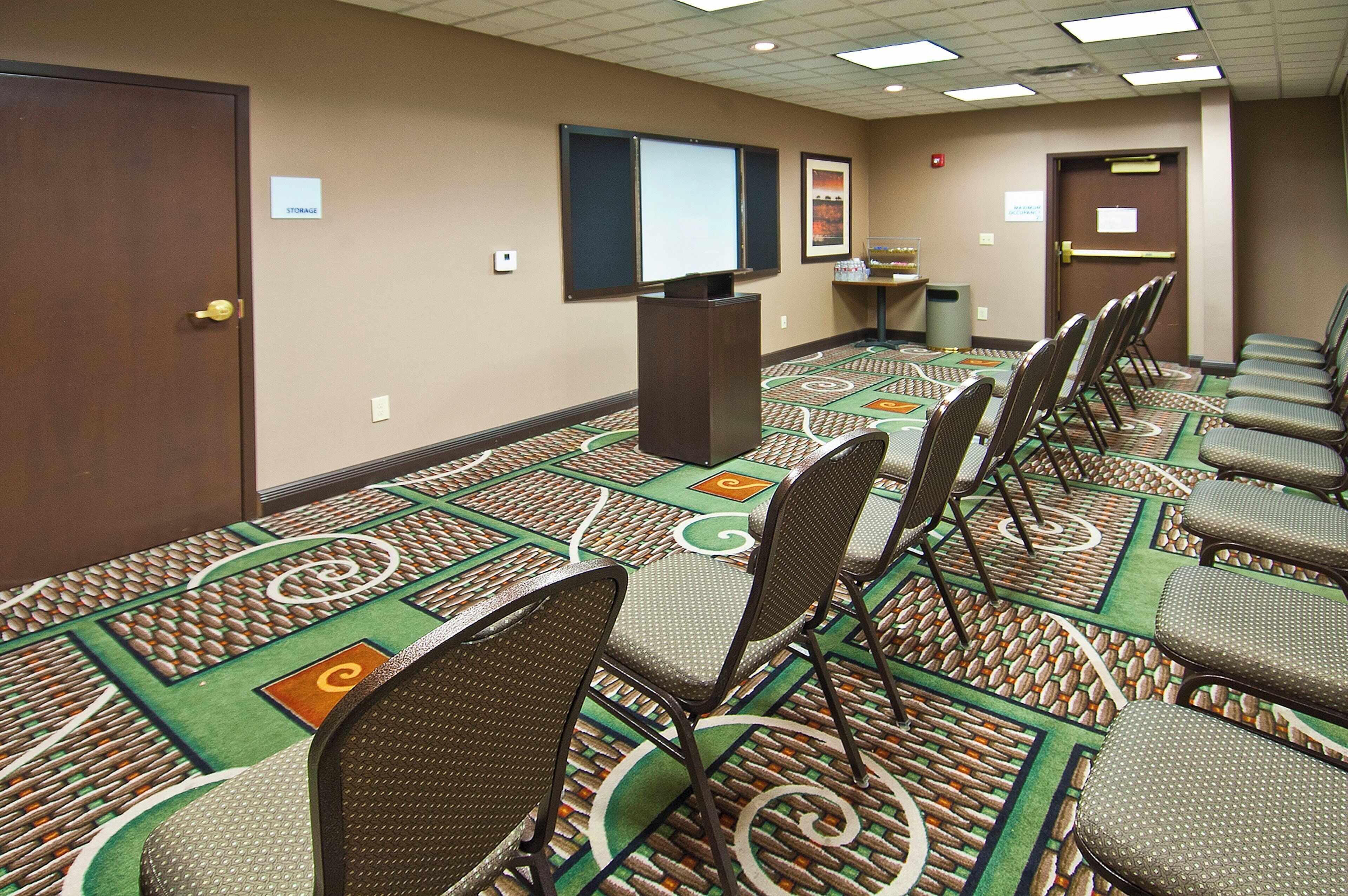 A conference room with a patterned carpet, rows of chairs, a podium, and a projector screen.
