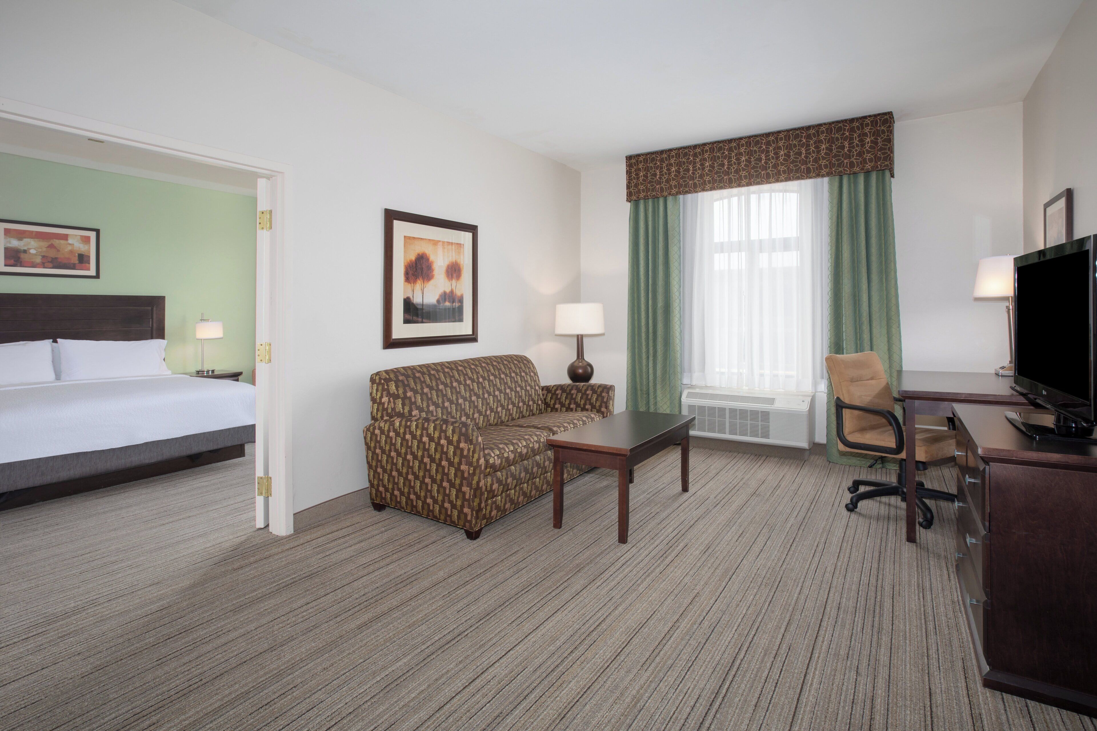 A hotel room with a bed, a sofa, a desk, a chair, a TV, and a window with curtains.