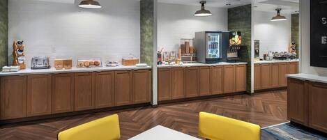 A modern kitchen with wooden cabinets, a refrigerator, and a coffee machine. There are two yellow chairs and a white table.