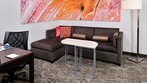 A hotel room with a sectional sofa, a small white table, a desk with a chair, a large abstract painting, and a floor lamp. - Springhill Suites by Marriott Oklahoma City Airport (Oklahoma City)
