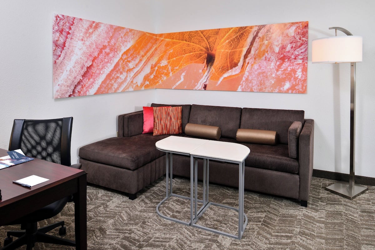 A hotel room with a sectional sofa, a small white table, a desk with a chair, a large abstract painting, and a floor lamp.