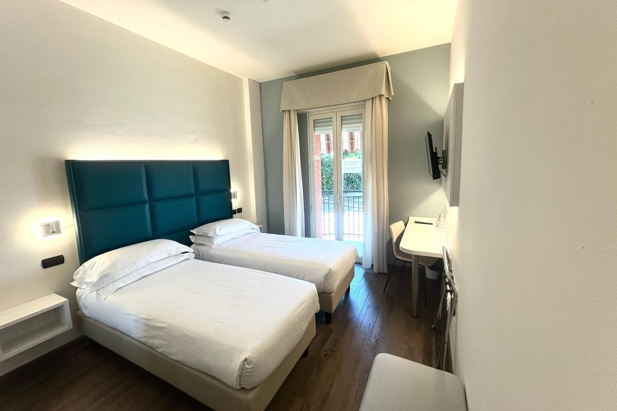 A hotel room with two beds, a small desk, a TV, and a balcony with curtains.