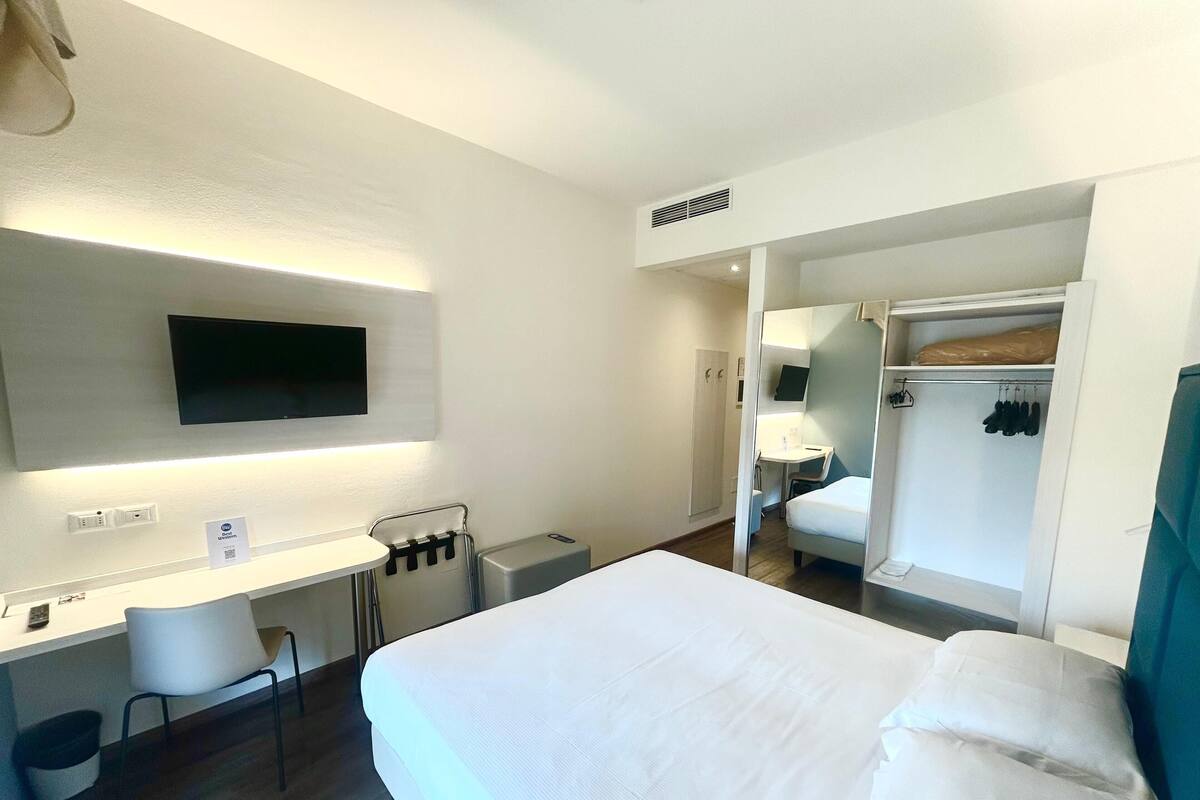 A hotel room with a bed, a desk, a chair, a TV, and a wardrobe.