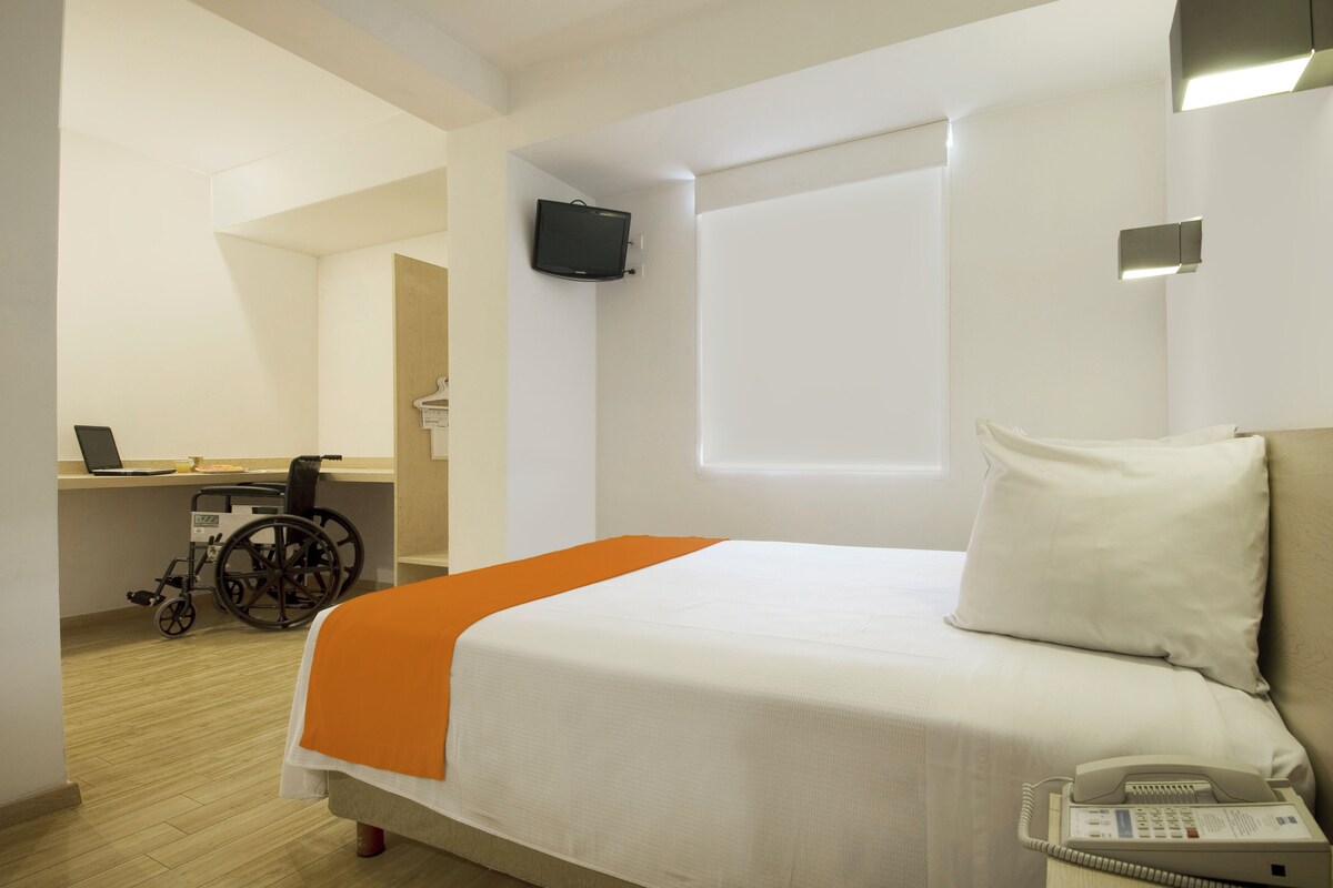 A hotel room with a bed, a desk, a wheelchair, and a TV mounted on the wall.